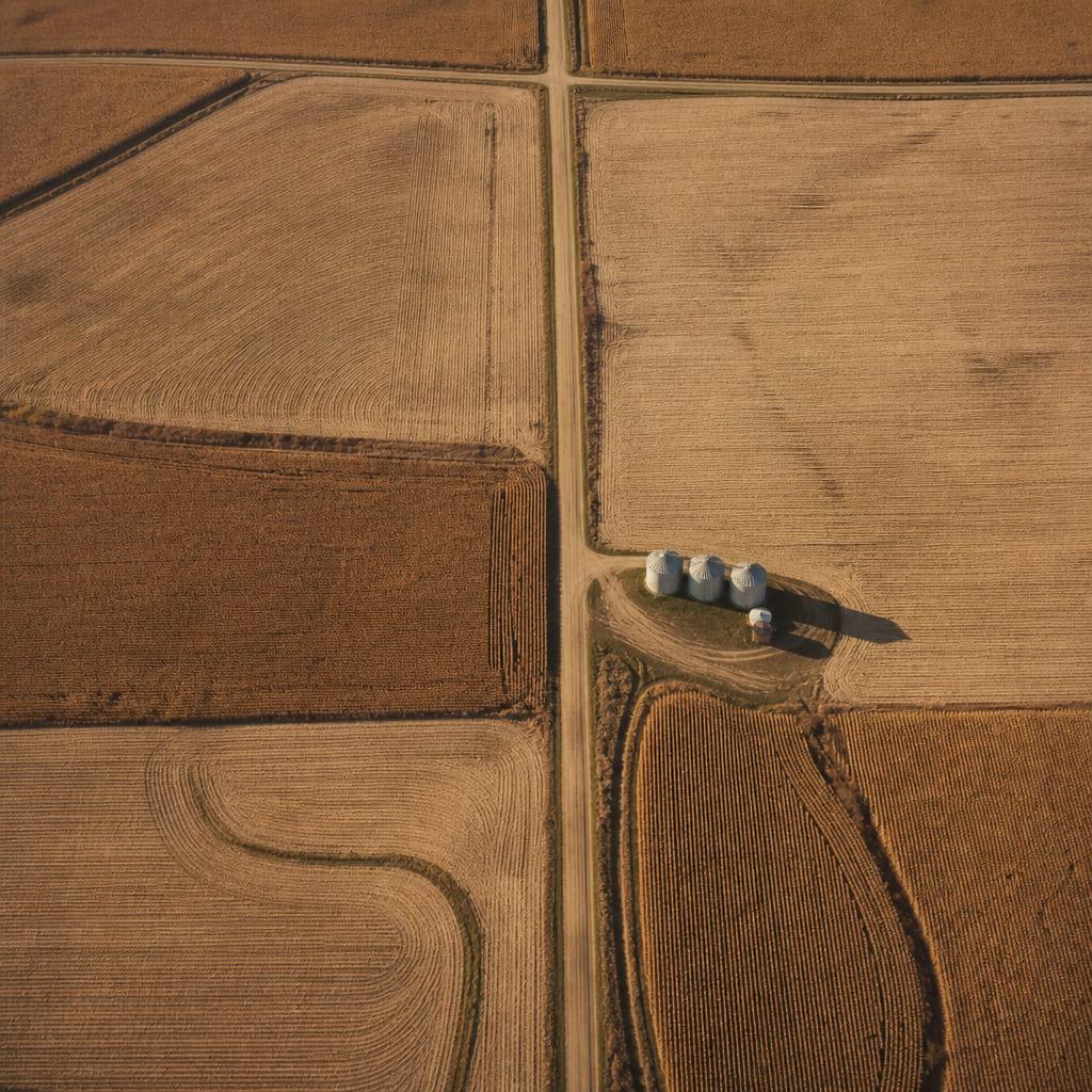 Aerial view of Midwestern row-crop farmland divided into a grid of fields, with grain bins beside a section road.